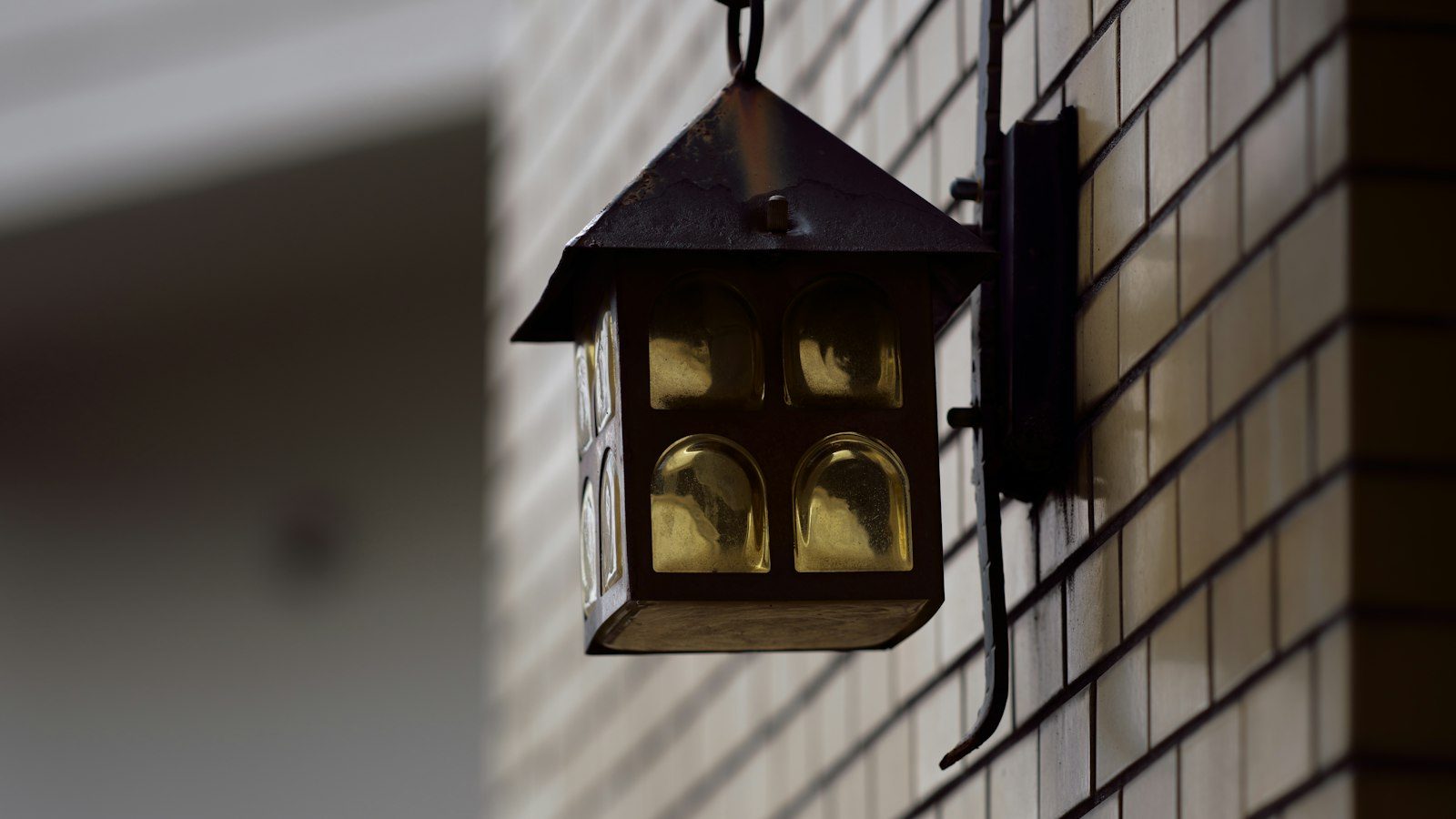 Square metal lantern with amber glass hanging from a wall hook beside a tiled facade