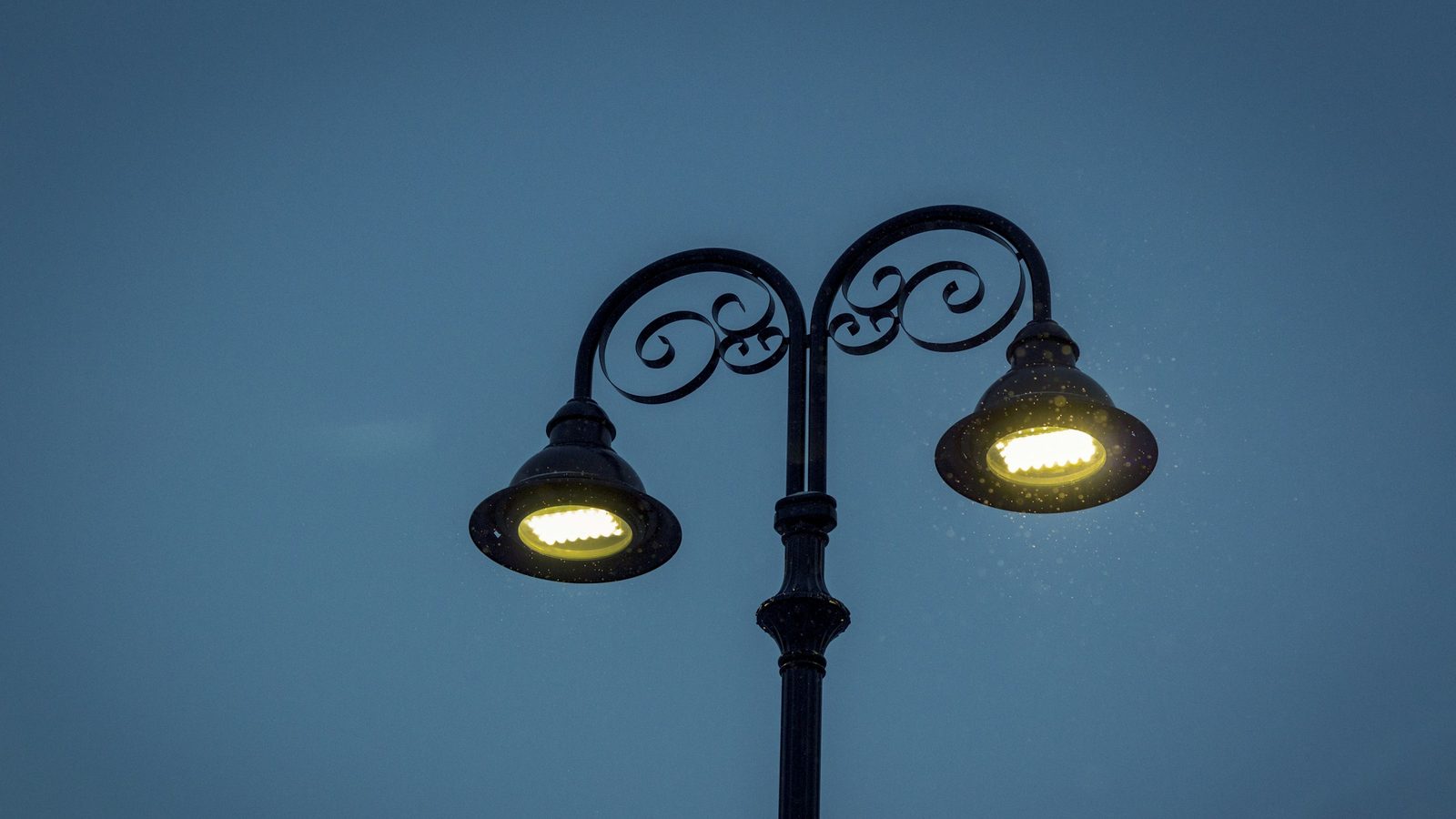 Black twin-arm lamp post with scrolled arms and two glowing bell-shaped heads against a dusk sky